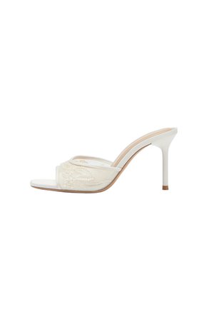 Mule blanche à talon haut avec bout ouvert et bride en dentelle transparente ornée de broderies florales et un talon aiguille fin.