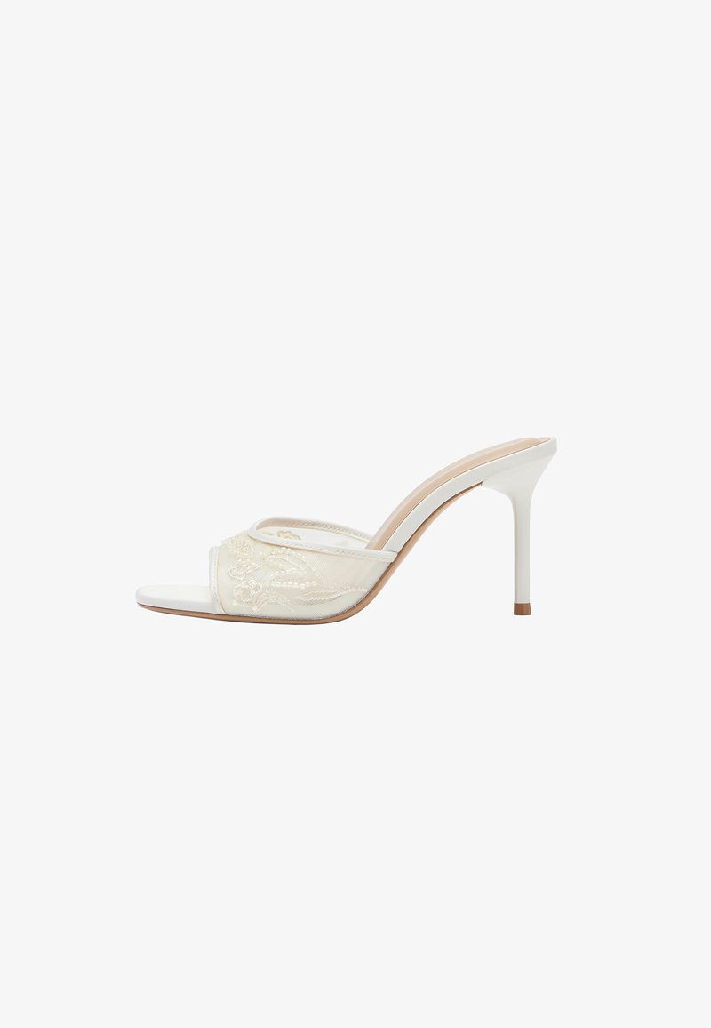 Mule blanche à talon haut avec bout ouvert et bride en dentelle transparente ornée de broderies florales et un talon aiguille fin.