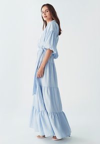 Světle modré dlouhé šaty s nabíranými rukávy, šňůrkou v pase a vrstveným volánkovým lemem. Vyrobené z jemné látky, předvádějící splývavou, uvolněnou siluetu.