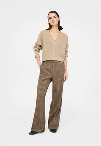 Beige neuleneuletakki, jossa on lyhyet hihat, V-pääntie ja napit. Yhdistetty ruskeisiin ja mustiin ruudullisiin leveälahkeisiin housuihin.