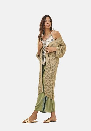 Larga chaqueta beige con patrón texturizado de diamantes, superpuesta a una camiseta de tirantes estampada y pantalones anchos verdes, combinada con sandalias planas doradas.