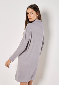 Robe en tricot côtelé gris avec un col montant, des manches longues et une coupe décontractée, tombant au milieu de la cuisse. Présente des fentes latérales pour faciliter les mouvements.