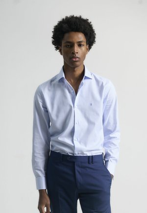 Camisa de vestir a rayas azul claro con collar abotonado, mangas largas y un pequeño emblema en el pecho, combinada con pantalones oscuros de color marino.