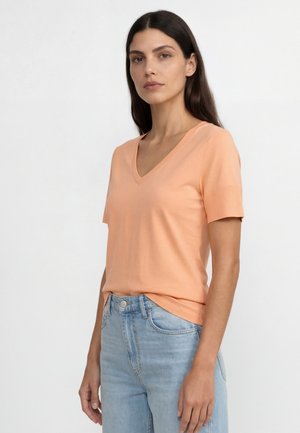 Donna con lunghi capelli scuri che indossa una maglietta pesca con scollo a V e maniche corte e jeans azzurri, in piedi davanti a uno sfondo bianco uniforme.