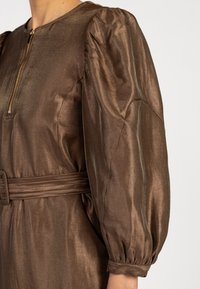 Robe longue en satin brun brillant à manches bouffantes, avec une ceinture à la taille et une fermeture éclair dorée au col.