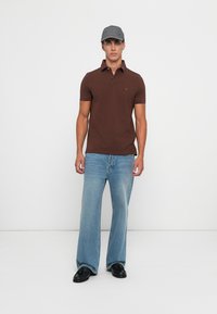 Mann som har på seg grå caps, brun kortermet polo-skjorte, lyseblå løse jeans og svarte sko, står vendt mot kameraet på hvit bakgrunn.