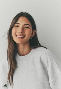 Grijze sweatshirt met een ronde hals, voorzien van een zachte textuur en een losse pasvorm. Lange donkere haren, subtiele hoepel oorbellen, en een neutrale achtergrond.