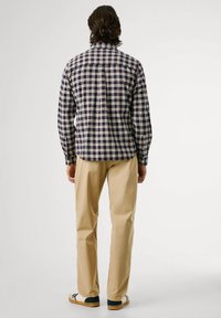 Uomo con capelli di lunghezza media che indossa una camicia a quadri navy, rossa e bianca a maniche lunghe, pantaloni beige e sneakers bianche, di spalle.