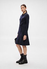 Tumši zilas kleita ar garām piedurknēm, apkakli un jostu viduklī. Mīksts audums, A formas siluets, papildināts ar melniem potīšu zābakiem un mazu somiņu.