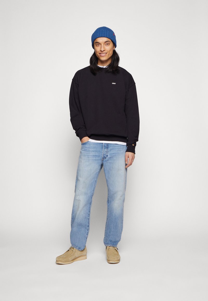 Schwarzer Sweatshirt mit Logo, hellblaue verwaschene Jeans und braune Schuhe. Getragen mit einer blauen gerippten Mütze. Lässiger Schnitt mit entspannter Silhouette.