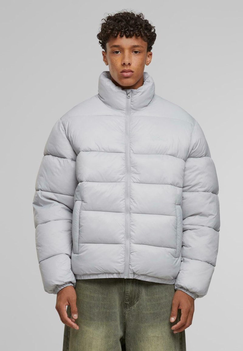 Jachetă puffer gri deschis, cu guler înalt, design matlasat, închidere cu fermoar pe față și manșete elastice. Textură netedă cu branding minim.