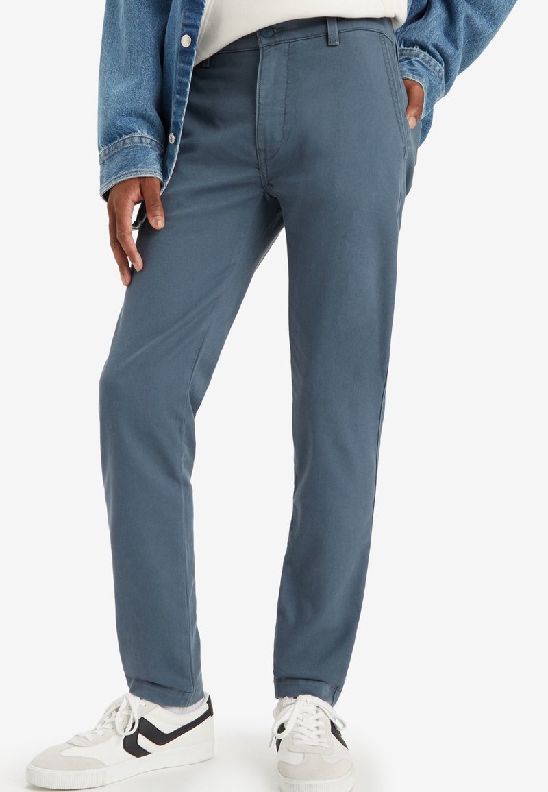 Pantalons chinos en coton bleu clair avec une coupe ajustée, passants de ceinture et poches latérales, associés à des sneakers blancs et noirs.