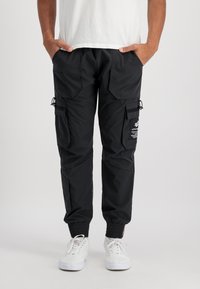 Person som bär svarta cargobyxor med flera fickor och elastiska manschetter, tillsammans med vita sneakers och en vit t-shirt.