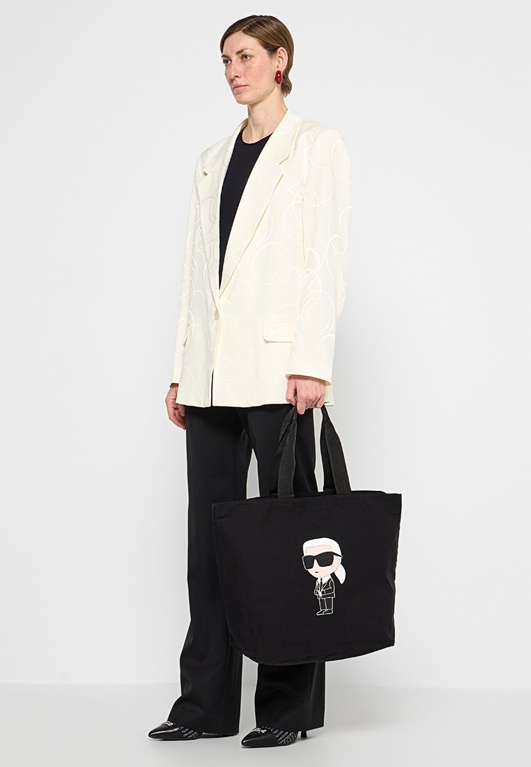 Schwarze Tragetasche mit einer Cartoonfigur in Sonnenbrille, die auf der Vorderseite aufgedruckt ist. Die Tasche hat stabile schwarze Tragegriffe und eine weiche Stoffstruktur.