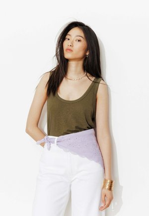 Femme aux longs cheveux foncés portant un débardeur vert olive, un pantalon blanc, une écharpe brodée lavande nouée autour de la taille et un bracelet manchette doré.