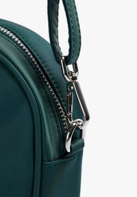 Esquina de una bolsa de nylon verde con un zipper plateado y detalles en metal. La correa presenta costuras texturizadas a lo largo del borde.