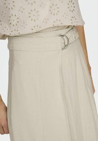 Beige Rock mit breitem Bund, der mit einem runden silbernen Schnallen-Detail versehen ist, und einer glatten Textur. Der Stoff hat vertikale Nähte für den stylischen Look.