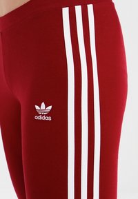 Röda träningsleggings i en bomullsblandning, med tre vita vertikala ränder och en broderad vit Adidas-logotyp.