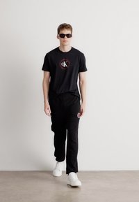 Μαύρο μπλουζάκι με λογότυπο, μαύρες φόρμες με έμβλημα, λευκά αθλητικά παπούτσια και γυαλιά ηλίου. Απλός σχεδιασμός, χαλαρή εφαρμογή και μονοχρωματική χρωματική παλέτα.