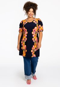 Marineblaues Tunika mit leuchtend pinkem und orangem Blumenmuster, Puffärmeln, kombiniert mit blauen Jeans und pinken Absatzsandalen, die einen geschichteten Look kreieren.