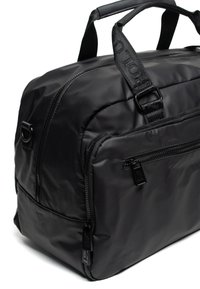 Černá taška typu duffel z nylonu s polstrovanou texturou, dvojitými horními rukojeťmi a několika kapsami na zip. Na popruzích jsou loga jako dekorace.