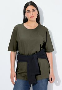 Olivgrünes kurzärmliges T-Shirt mit lässiger Passform, kombiniert mit einem dunkelblauen Pullover, der um die Taille gebunden ist; schlichte Textur, runder Ausschnitt.