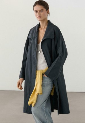 Donna che indossa un lungo cappotto scuro sopra una canotta grigia con un maglione giallo legato in vita e jeans azzurri.