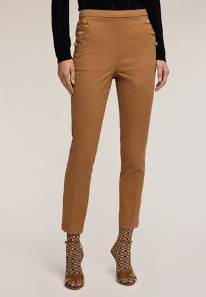 Pantalones ajustados color beige con una textura suave, con acentos de botones dorados a los lados. Combinados con calcetas de red negra con patrón y tacones.