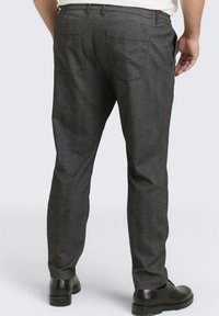 Man wearing dark gray dress pants and black dress shoes, shown from the back against a plain light background.