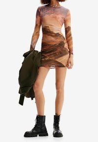 Robe à manches longues avec imprimé paysage, design froncé aux tons chauds ; associée à une veste vert olive et des bottes de combat noires.