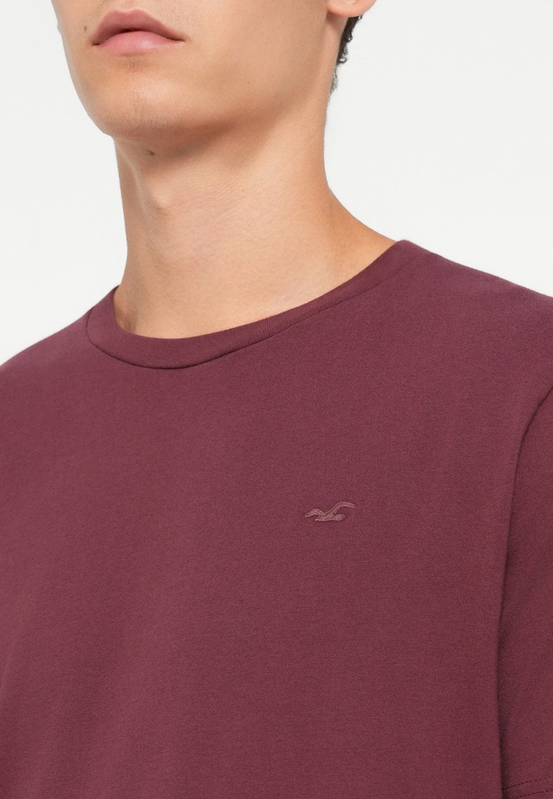 Primo piano di una persona che indossa una semplice maglietta a girocollo color mogano con un piccolo logo ricamato sul petto.