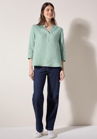 Top in lino verde chiaro a maniche lunghe con scollo a V e vestibilità ampia, abbinato a jeans in denim blu scuro a gamba larga e sneakers bianche.