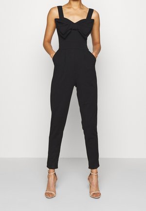 Combinaison noire avec larges bretelles, grand nœud avant, corsage ajusté, jambes fuselées et poches latérales, réalisée en tissu doux.