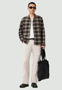 Homem a usar óculos de sol pretos, camisa xadrez castanha, t-shirt branca, calças bege, cinto preto, sapatos pretos, segurando uma bolsa tote preta.