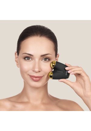 Femme tenant un appareil de massage facial noir et doré sur sa joue droite, souriant sur un fond neutre.