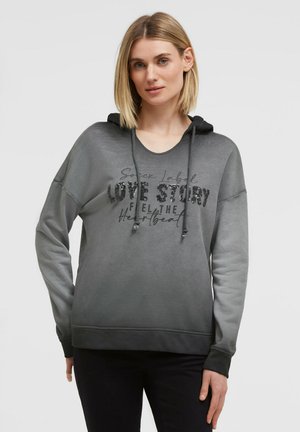 Woman with short blonde hair wearing a gray hooded sweatshirt with "Love Story" text in black sequins on the front.