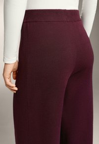 Pantaloni tricotati burgundy cu un brâu lat, având o textură netedă și accente cu nervuri pe laturi pentru un detaliu suplimentar.