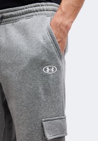 Άτομο που φοράει γκρι φόρμα Under Armour με το χέρι στην πλαϊνή τσέπη και μια τσέπη με καπάκι στο μηρό, σε μονόχρωμο φόντο.