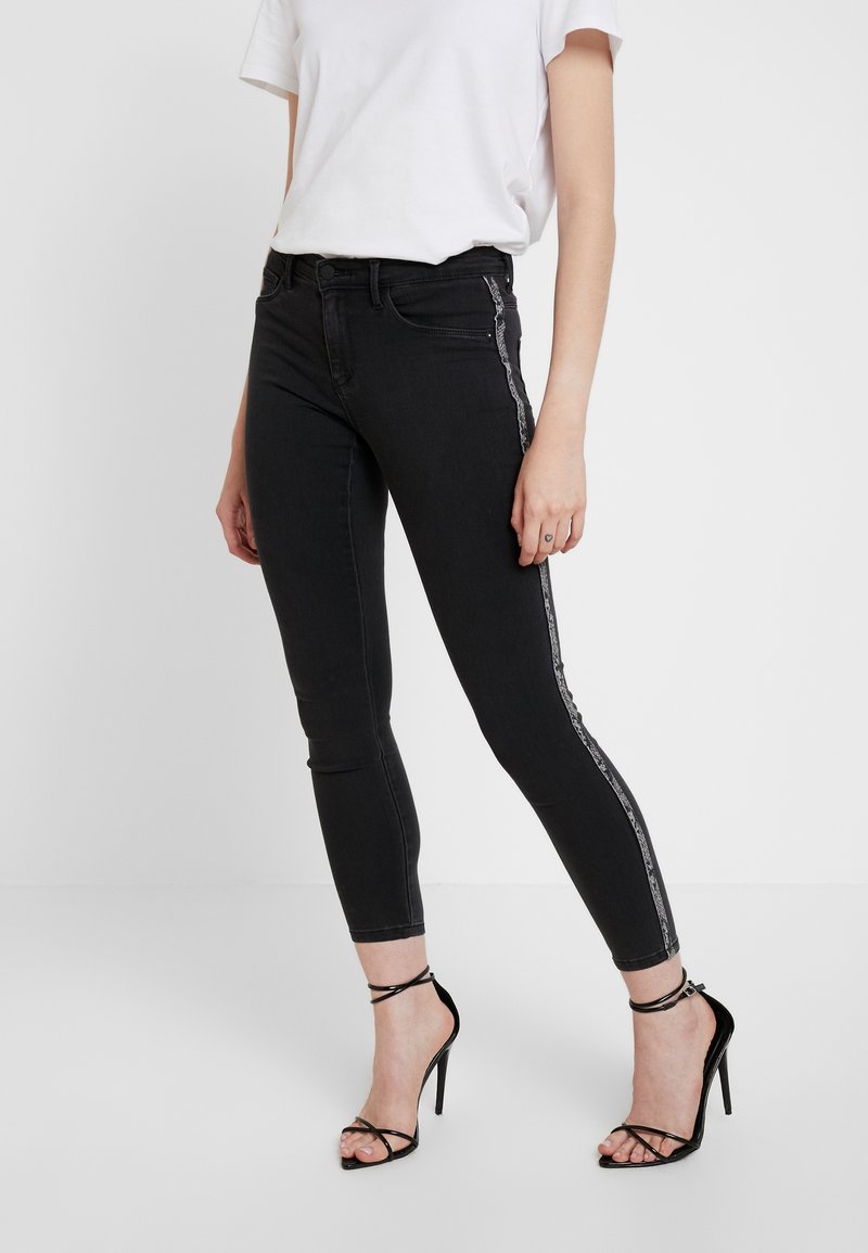 Femme portant un jean skinny noir avec des bandes latérales argentées, un t-shirt blanc et des sandales noires à talons hauts et à brides, se tenant devant un fond blanc.