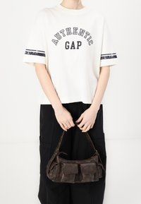 Bílý tričko s potiskem "AUTENTICKÝ GAP", v kombinaci s černými širokými kalhotami a hnědou koženou kabelkou s předními kapsami a krátkým uchem.