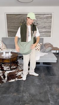 Grüne Umhängetasche aus weichem, strukturiertem Stoff, quer getragen. Model in einem gestuften Outfit: einer grünen Weste über einem weißen T-Shirt und beigen Hosen.