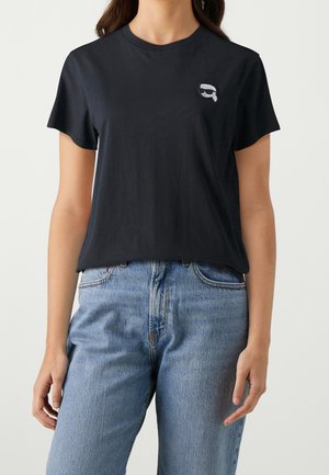 Nainen, jolla on päällään musta lyhythihainen t-paita, jossa on pieni harmaa kuvio vasemmassa rinnassa, sekä vaaleansiniset korkeavyötäröiset farkut.