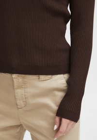 Maglione a costine marrone con maniche lunghe, tessuto testurizzato e design aderente, abbinato a pantaloni beige chiaro.