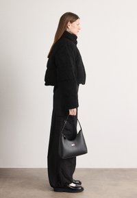 Svart kort faux fur-jakke, svarte vide bukser og en svart håndveske med en liten logo. Modellen har på seg svarte sko og sølvringer.