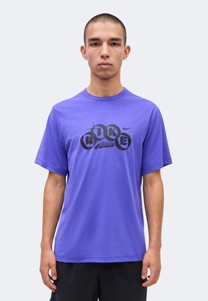 Tânăr cu păr scurt purtând un tricou Nike violet cu text grafic negru, stând cu brațele relaxate pe un fundal simplu.