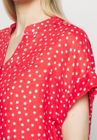 Czerwona bluzka z krótkim rękawem wykonana z lekkiego materiału, zdobiona białymi kropkami, z dekoltem w kształcie litery V i wywiniętymi mankietami.