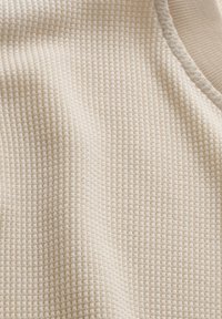 Nahaufnahme eines cremefarbenen Waffelstrickstoffs mit geripptem Abschluss, der die Textur und Details des Strickmusters zeigt.