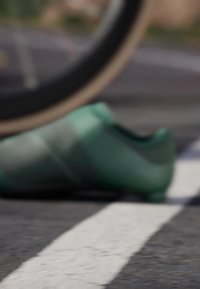 Green athletic shoe with a textured surface and angular design, partially obscured by a bicycle wheel, resting on a pavement with white lines.