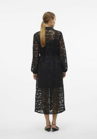 Abito in pizzo nero con un motivo floreale trasparente. Presenta maniche lunghe, un taglio a metà lunghezza e un dettaglio in vita con lacci.