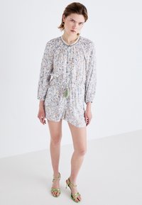 Blomstrete romper i lett stoff, med lange ermer, snøring i midjen og dusker, parret med grønne sandaletter med stropper.